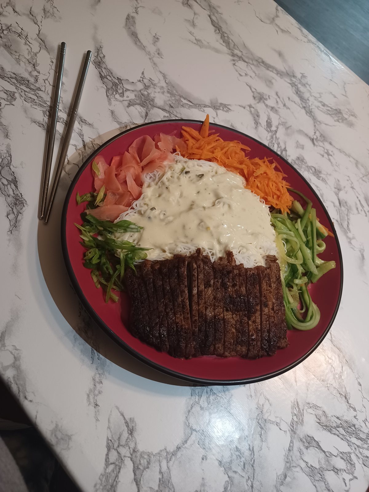 Sliced blackened steak over noodles with creamy sauce, carrots, cucumber and pickled ginger